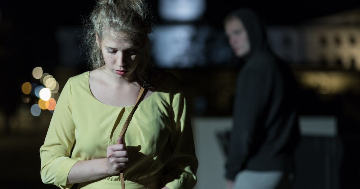 Blonde woman wearing a yellow shirt walking alone at night with a nefarious looking man behind her.